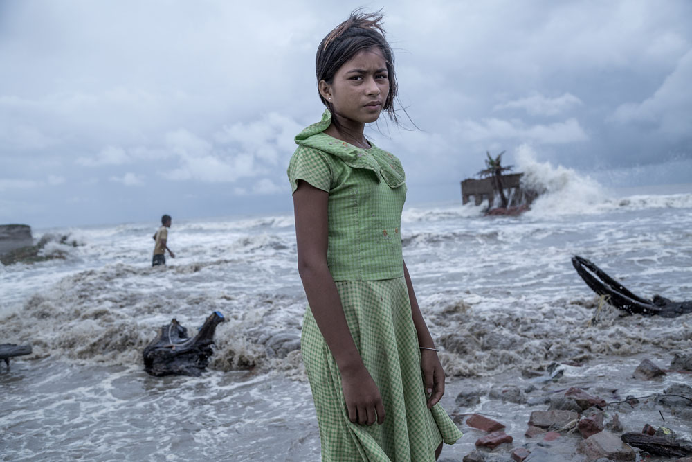 18. August 2020: Ein Mädchen, das auf der Insel Namkhana vor ihrem völlig zerstörtem Teeladen steht. Die als Weltkulturerbe bekannte Region zwischen Indien und Bangladesch gelegen heiß Sundarbans (bengalisch für "schöner Wald) und ist bekannt für ihre enormen Waldressourcen. Diese bilden die Lebensgrundlage der Einheimischen. In den letzten Jahren wurde die globale Erwärmung jedoch zu einem zunehmenden Problem. Durch das wahllose Abholzen von Bäumen für den gewerblichen und privaten Gebrauch entstand ein Ungleichgewicht, das das Land schwächte, das häufig Opfer von Stürmen, starken Regenfällen und Überschwemmungen wurde. Jedes Jahr steigt der Meeresspiegel um ungefähr 3,14 mm, wodurch bereits mehrere Inseln unter Wasser liegen. Erhöhte Bodenerosion, Verlust der Bodenqualität, die zu minimierten Ernteerträgen führt, haben die Situation noch verschlimmert. Das Wasser hat bereits Teile von Mousuni und Ghoramara Island verschlungen. Wenn die dieser Trend weiter anhält, wird ein Großteil der Sundarbans bald im Wasser versunken sein. (c) Supratim Bhattacharjee, Indien